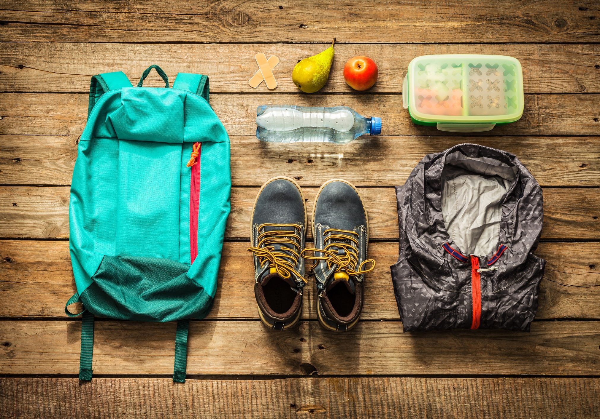 Traveling - packing (preparing) for adventure school trip concept. Backpack, boots, jacket, lunch box, water and fruits on wooden background captured from above (flat lay).