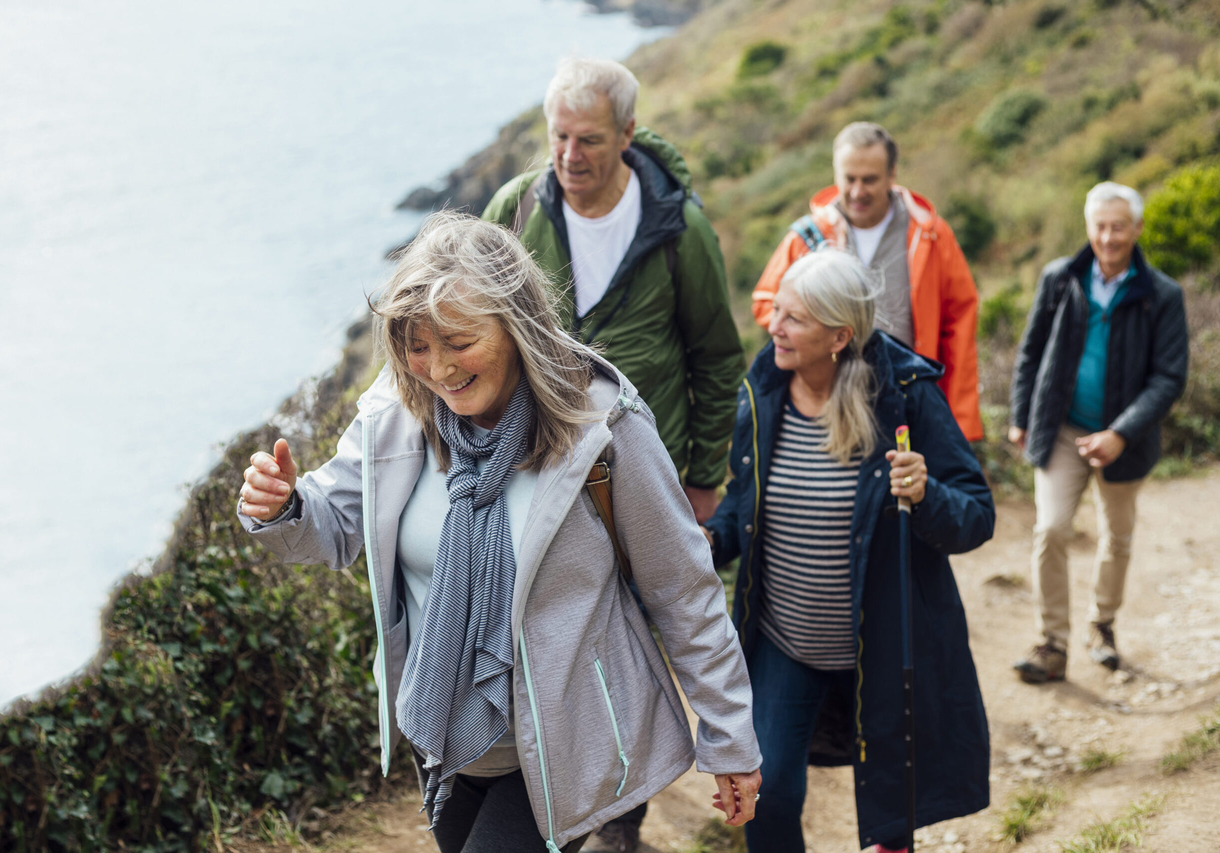 A group of male and female friends walking up a hill on a coastal path along a cliff edge overlooking the sea in Cornwall. The main focus is one woman wearing a jacket and scarf, she is smiling while looking down.
